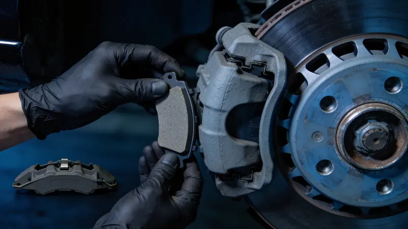 Gloved hands holding a brake pad beside a brake caliper and rotor, wheel off
