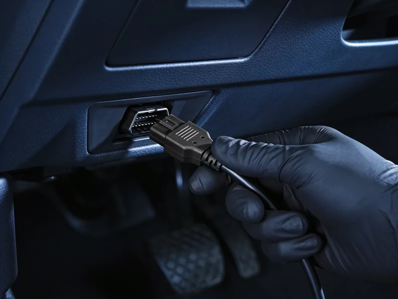 A gloved hand plugging a diagnostic connector into the port under a dashboard